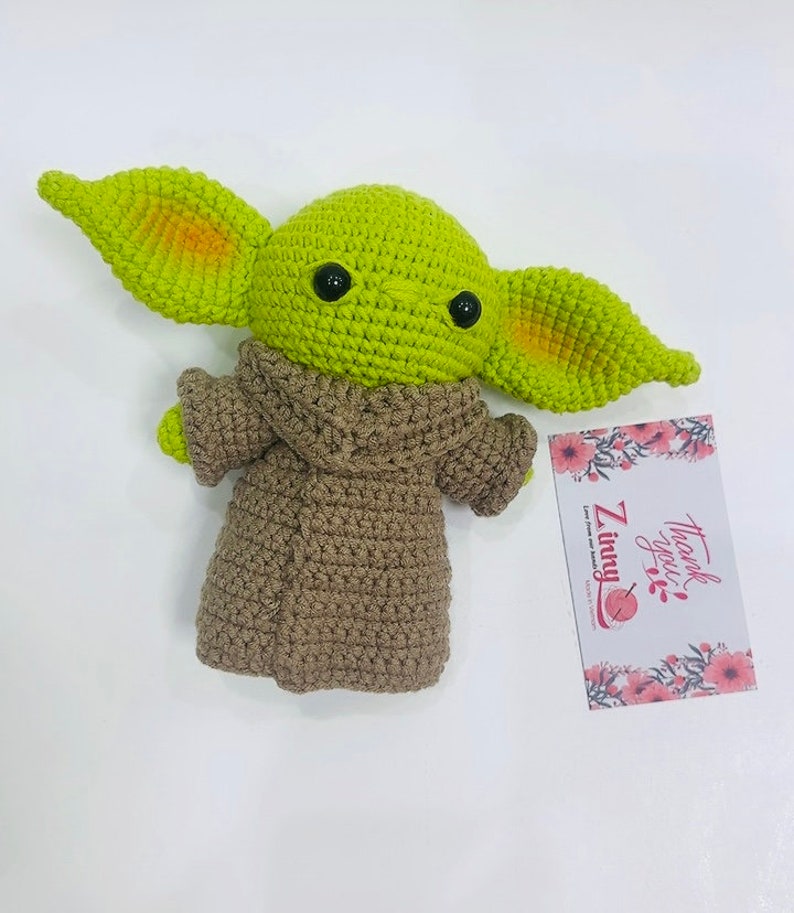 May include: Green crocheted plush toy with large ears and brown crocheted clothing. The toy is sitting on a white surface with a thank you card.
