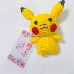 Könnte beinhalten: Ein gelbes, gehäkeltes Pikachu-Plüschtier mit schwarzen Augen, roten Wangen und einer schwarzen Nase. Das Spielzeug sitzt auf einer weißen Oberfläche mit einer rosa-weißen Blumenkarte, auf der "Danke" und "Zinnny" mit einem Wollknäuel-Symbol steht.