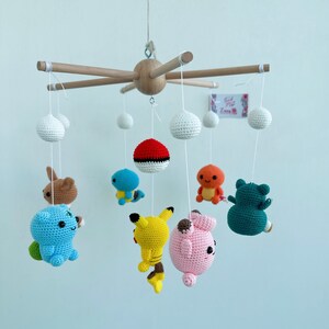 May include: A wooden mobile with crocheted Pokemon characters hanging from it. The characters include Pikachu, Bulbasaur, Charmander, Squirtle, Jigglypuff, and a Pokeball. The mobile is white and has a wooden base.