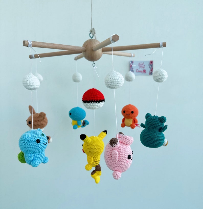 May include: A wooden mobile with crocheted Pokemon characters hanging from it. The characters include Pikachu, Bulbasaur, Charmander, Squirtle, Jigglypuff, and a Pokeball. The mobile is white and has a wooden base.