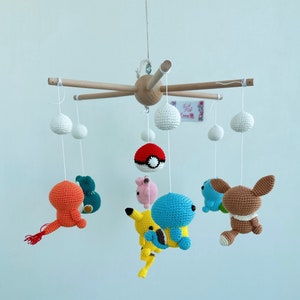 May include: A wooden baby mobile with crocheted characters from the Pokemon franchise. The mobile features Pikachu, Squirtle, Bulbasaur, Charmander, and a Pokeball. The mobile is suspended from a wooden base with white strings.