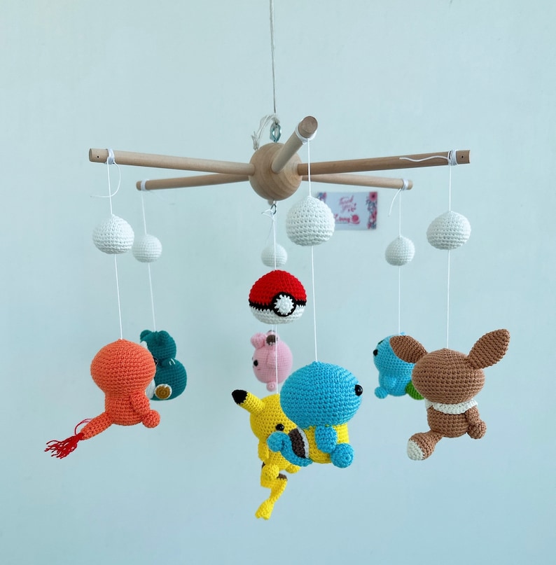 May include: A wooden baby mobile with crocheted characters from the Pokemon franchise. The mobile features Pikachu, Squirtle, Bulbasaur, Charmander, and a Pokeball. The mobile is suspended from a wooden base with white strings.