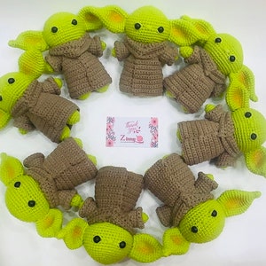 May include: A group of nine crocheted green and brown plush toys resembling the character Baby Yoda from the Star Wars series. The toys are arranged in a circle on a white surface.