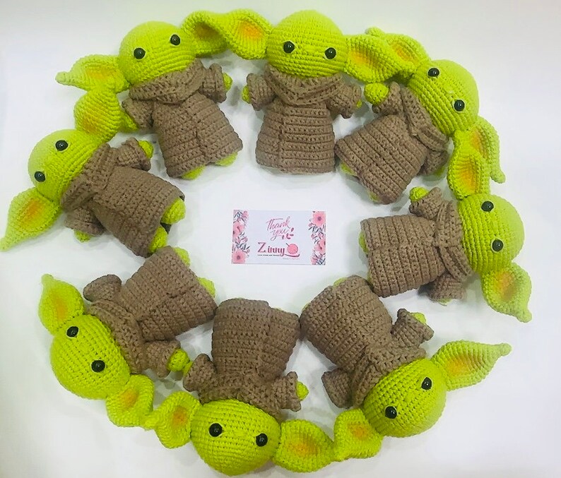 May include: A group of nine crocheted green and brown plush toys resembling the character Baby Yoda from the Star Wars series. The toys are arranged in a circle on a white surface.