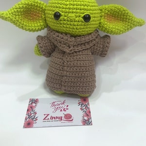 May include: A green crocheted plush toy with large ears and brown crocheted clothing. The toy is sitting on a white surface with a small card that says "Thank you" and "Z inny" with a small crocheted yarn ball.