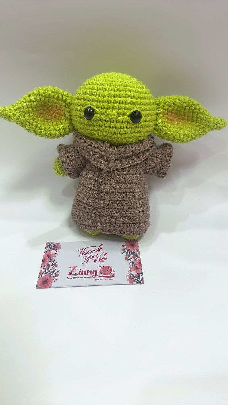 May include: A green crocheted plush toy with large ears and brown crocheted clothing. The toy is sitting on a white surface with a small card that says "Thank you" and "Z inny" with a small crocheted yarn ball.