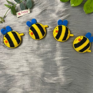 Peut inclure: Quatre jouets en forme d'abeille en crochet avec des rayures jaunes et noires, des ailes bleues et des visages orange. Les abeilles sont sur une surface grise et duveteuse.