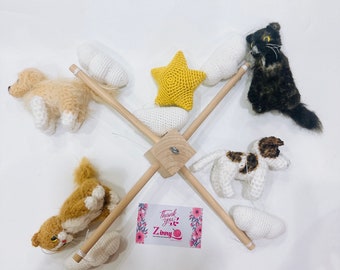 Op de afbeelding: Een houten babymobiel met gehaakte dieren en wolken. De mobiel heeft een bruine en witte hond, een bruine en witte kat, een zwart-witte kat, een gele ster en witte wolken. De mobiel hangt aan een houten frame.