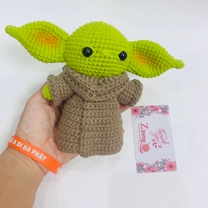 May include: Green crocheted plush toy with large ears and brown crocheted coat. The toy has black eyes and a small mouth. The toy is being held in a hand. The background is white.