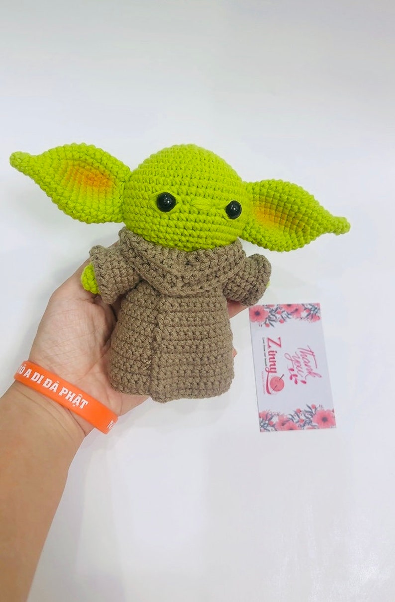 May include: Green crocheted plush toy with large ears and brown crocheted coat. The toy has black eyes and a small mouth. The toy is being held in a hand. The background is white.