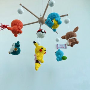 May include: A wooden mobile with crocheted characters hanging from it. The characters include a yellow Pikachu, a blue Squirtle, a brown Eevee, a red and orange Charmander, a green Bulbasaur, and a blue and white Pokeball.