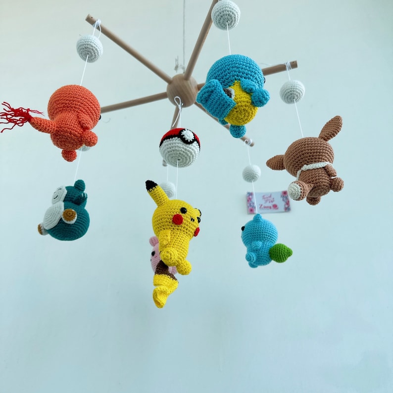 May include: A wooden mobile with crocheted characters hanging from it. The characters include a yellow Pikachu, a blue Squirtle, a brown Eevee, a red and orange Charmander, a green Bulbasaur, and a blue and white Pokeball.