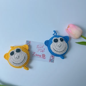 Può includere: Due ornamenti a forma di scimmia all'uncinetto. Uno è giallo con un viso beige e occhi neri, l'altro è blu con un viso beige e occhi neri. Una carta con il testo "Thank you" e il marchio "Zinny" è tra loro. Un tulipano rosa è sullo sfondo.
