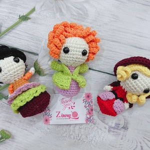 May include: Three crocheted dolls with different colored outfits and hair. The doll on the left is wearing a green and burgundy dress. The doll in the middle is wearing a green and purple dress with orange hair. The doll on the right is wearing a red and purple dress with a burgundy hood. The dolls are sitting on a wooden surface with a thank you card in the background.