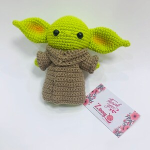 May include: A green crocheted plush toy with large ears and brown crocheted clothing. The toy has black button eyes and is sitting on a white surface. The text "Thank you" and "Z Tiny" is visible on a card in the background.