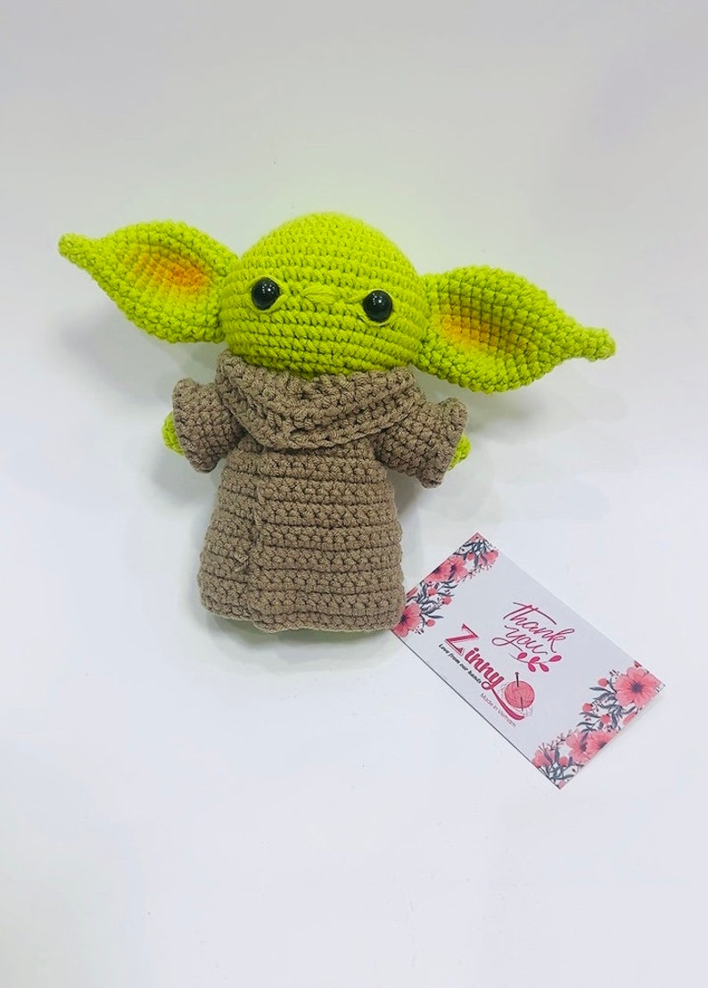May include: A green crocheted plush toy with large ears and brown crocheted clothing. The toy has black button eyes and is sitting on a white surface. The text "Thank you" and "Z Tiny" is visible on a card in the background.