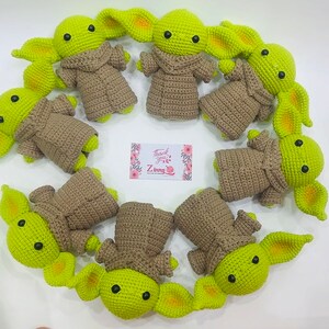 May include: A collection of nine crocheted green and brown baby Yoda plushies. The plushies are arranged in a circle on a white surface. The plushies have large ears and big eyes.