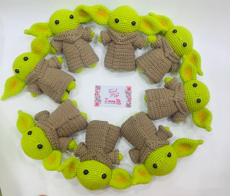 May include: A collection of nine crocheted green and brown baby Yoda plushies. The plushies are arranged in a circle on a white surface. The plushies have large ears and big eyes.