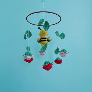 May include: A handmade crochet baby mobile featuring a yellow and black bee, green leaves, and red and pink flowers. The mobile is suspended from a brown ring and hangs against a light blue background.