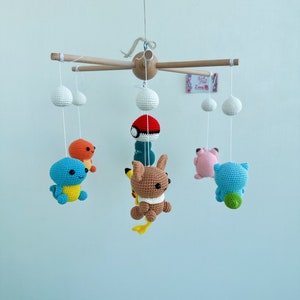 May include: A wooden mobile with crocheted Pokemon characters hanging from it. The characters include Bulbasaur, Charmander, Squirtle, Pikachu, and a pink Pokemon. The mobile is suspended from a wooden dowel rod.