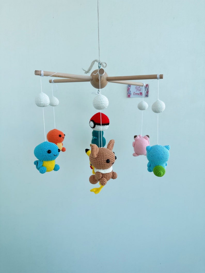 May include: A wooden mobile with crocheted Pokemon characters hanging from it. The characters include Bulbasaur, Charmander, Squirtle, Pikachu, and a pink Pokemon. The mobile is suspended from a wooden dowel rod.