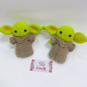 May include: Two crocheted green and brown plush toys resembling the character Baby Yoda from the Star Wars series. The toys are wearing brown robes and have large ears.