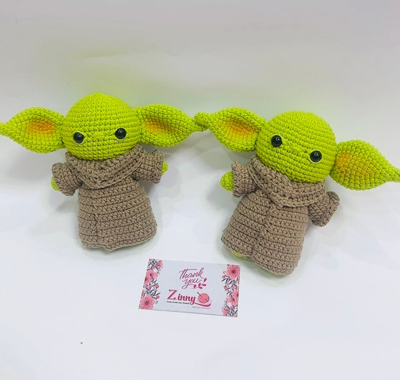 May include: Two crocheted green and brown plush toys resembling the character Baby Yoda from the Star Wars series. The toys are wearing brown robes and have large ears.