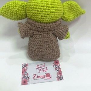 May include: A crocheted green and brown plush toy resembling a character from the Star Wars franchise. The toy is wearing a brown robe and has large ears. The toy is sitting on a white surface with a small card that says "Thank you" and "Zinny" with the text "Love from our hands" and "Made in Vietnam".