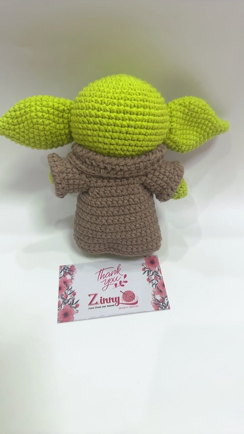 May include: A crocheted green and brown plush toy resembling a character from the Star Wars franchise. The toy is wearing a brown robe and has large ears. The toy is sitting on a white surface with a small card that says "Thank you" and "Zinny" with the text "Love from our hands" and "Made in Vietnam".