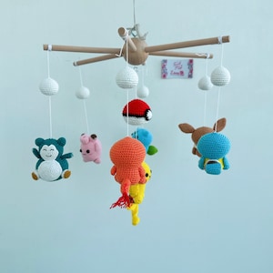 May include: A wooden mobile with crocheted Pokemon characters hanging from it. The characters include Bulbasaur, Charmander, Squirtle, Pikachu, and a Pokeball. The mobile is suspended from a wooden frame.