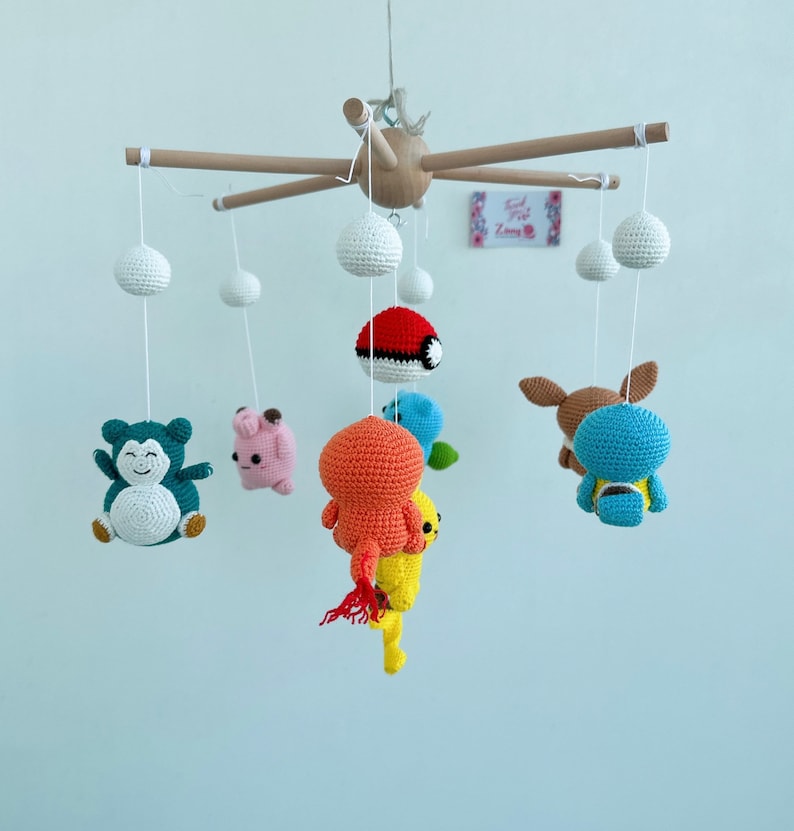 May include: A wooden mobile with crocheted Pokemon characters hanging from it. The characters include Bulbasaur, Charmander, Squirtle, Pikachu, and a Pokeball. The mobile is suspended from a wooden frame.