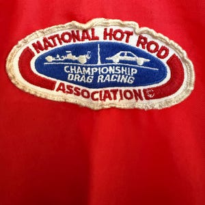 May include: Red fabric featuring an embroidered patch with the text "NATIONAL HOT ROD CHAMPIONSHIP DRAG RACING ASSOCIATION". The patch has a white border and car illustrations.