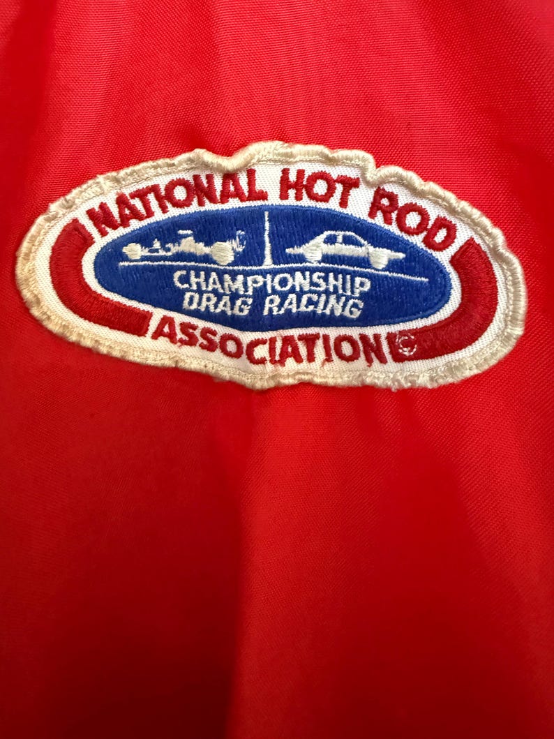May include: Red fabric featuring an embroidered patch with the text "NATIONAL HOT ROD CHAMPIONSHIP DRAG RACING ASSOCIATION". The patch has a white border and car illustrations.