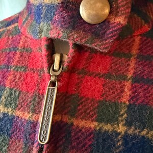 Puede incluir: Primer plano de una chaqueta a cuadros rojos, azules y verdes. La chaqueta tiene un tirador de cremallera de lat&oacute;n con la palabra "PENDLETON" grabada. Se ve un bot&oacute;n grande y redondo de lat&oacute;n cerca del cuello.