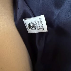 Puede incluir: Primer plano del interior de una prenda azul marino, que revela una etiqueta blanca. La etiqueta muestra el logotipo de Woolmark, junto con el texto "100% Pure New Wool" y un n&uacute;mero de serie. La tela parece suave y lujosa.