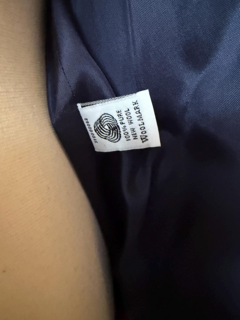 Puede incluir: Primer plano del interior de una prenda azul marino, que revela una etiqueta blanca. La etiqueta muestra el logotipo de Woolmark, junto con el texto "100% Pure New Wool" y un n&uacute;mero de serie. La tela parece suave y lujosa.