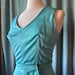 May include: A sleeveless turquoise dress featuring a V-neckline, vertical seams along the bodice, and a matching fabric tie belt at the waist. The garment is displayed on a mannequin, showcasing a smooth, solid-colored fabric with a slight sheen.