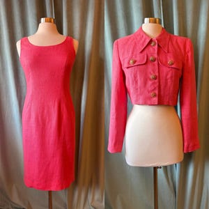 May include: A sleeveless, knee-length, coral-pink dress and a matching cropped jacket with gold-colored buttons. The dress has a scoop neckline. The jacket has long sleeves and a collared neckline. Both garments are displayed on mannequins.