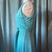 May include: A sleeveless turquoise dress featuring a fitted bodice with a side-tie waist detail and a flowing skirt. The garment is displayed on a mannequin, showcasing a smooth, slightly lustrous fabric texture and vertical seam construction.