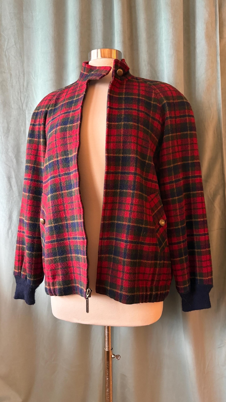 Puede incluir: Chaqueta a cuadros rojos, verdes y azul marino con cremallera y cuello alzado. La chaqueta tiene pu&ntilde;os de canal&eacute; azul marino y botones dorados. La chaqueta se exhibe en un maniqu&iacute;.