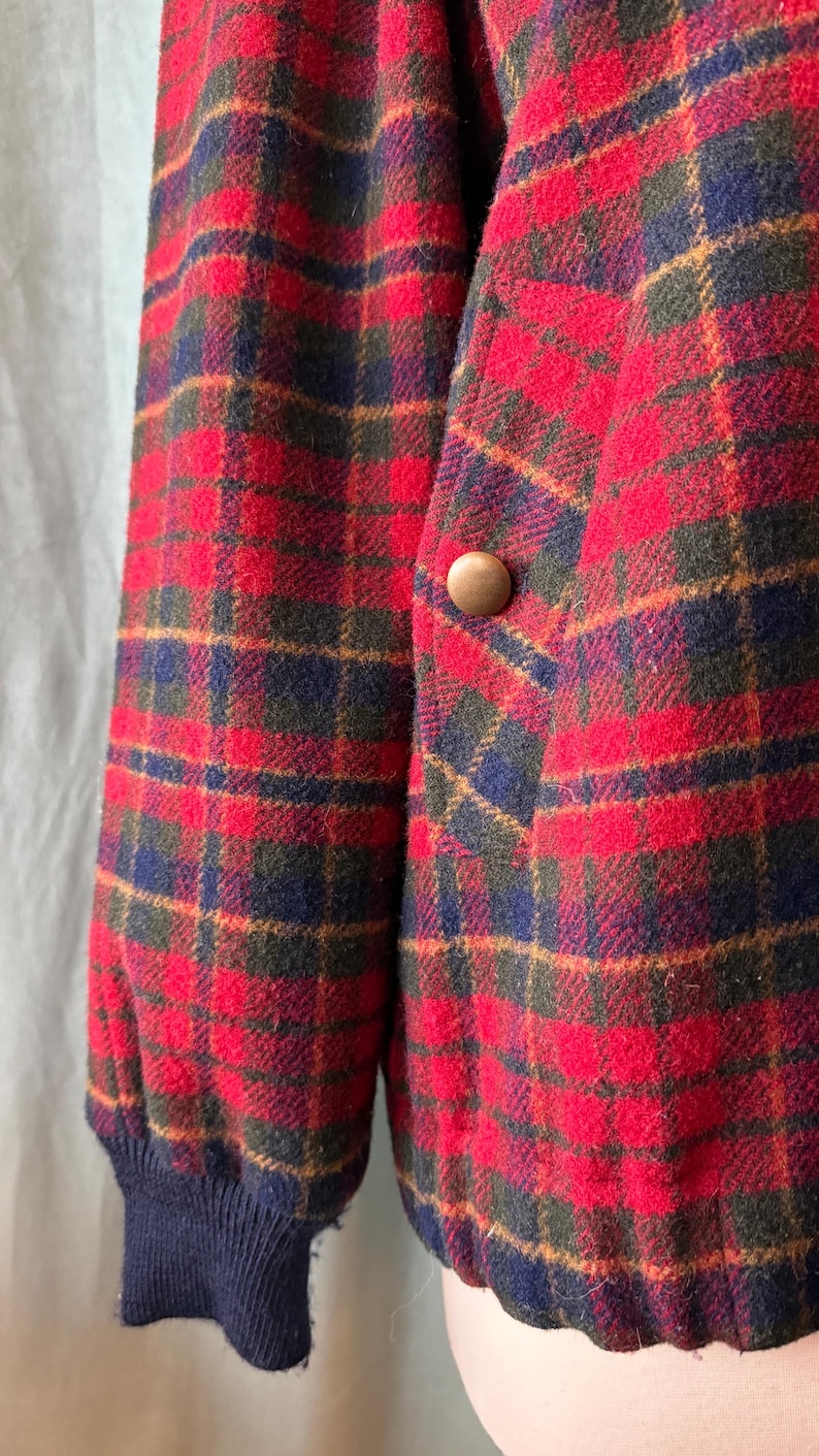 Puede incluir: Primer plano de una chaqueta a cuadros roja, azul y verde. La chaqueta tiene un bot&oacute;n de madera y un pu&ntilde;o azul marino acanalado. La tela tiene un aspecto texturizado. La chaqueta es de estilo cl&aacute;sico.