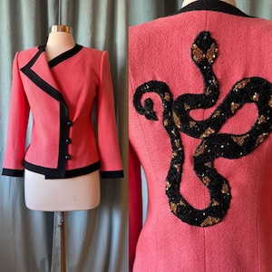 May include: A coral-colored blazer with black trim and buttons. The back of the jacket features a large, sequined snake design in black and gold. The jacket is displayed on a mannequin.