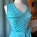 May include: A sleeveless turquoise dress featuring a V-neckline, a fitted bodice with vertical seam detailing, and a wide integrated waistband that ties at the side. The garment is displayed on a mannequin, showcasing a smooth, solid-colored fabric.