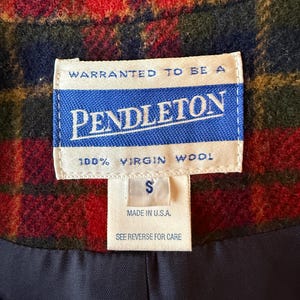 Puede incluir: Primer plano de una prenda de lana Pendleton con un estampado de cuadros rojos, verdes y azules. La etiqueta dice "PENDLETON 100% VIRGIN WOOL" y "MADE IN U.S.A." con una etiqueta de talla "S". El forro es azul marino.