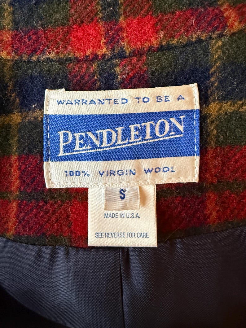 Puede incluir: Primer plano de una prenda de lana Pendleton con un estampado de cuadros rojos, verdes y azules. La etiqueta dice "PENDLETON 100% VIRGIN WOOL" y "MADE IN U.S.A." con una etiqueta de talla "S". El forro es azul marino.