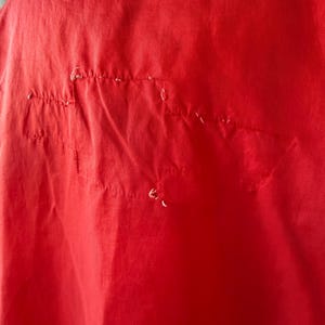 May include: Close-up of a vibrant red fabric showing stitching and minor damage. The material is a solid colour, potentially part of a garment or textile.