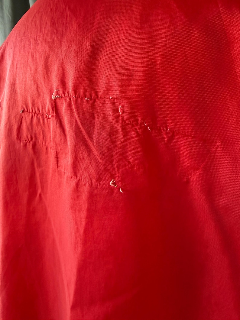 May include: Close-up of a vibrant red fabric showing stitching and minor damage. The material is a solid colour, potentially part of a garment or textile.