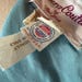 May include: A light blue garment featuring a white fabric label with the text Futura Couture of New York in magenta script. A smaller white tag displays the ILGWU union logo in red and blue with the text Made in U.S.A. and a care instruction tag.