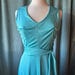 May include: A sleeveless turquoise dress featuring a V-neckline and a matching fabric tie-waist belt. The garment is constructed from a smooth, slightly shimmering material that drapes from the bodice down to the skirt, creating a clean silhouette.