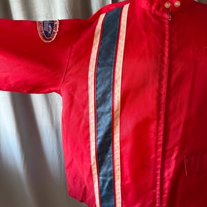 May include: A red racing jacket featuring a central blue stripe flanked by white stripes. The jacket has a high collar, a zip, and a patch on the sleeve with text. The jacket is a vintage style.
